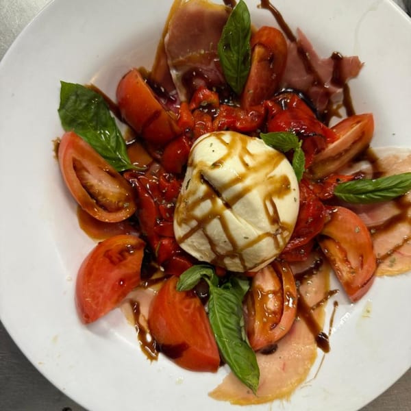A white plate of Caprese salad with burrata, tomatoes, basil, prosciutto, and balsamic glaze.