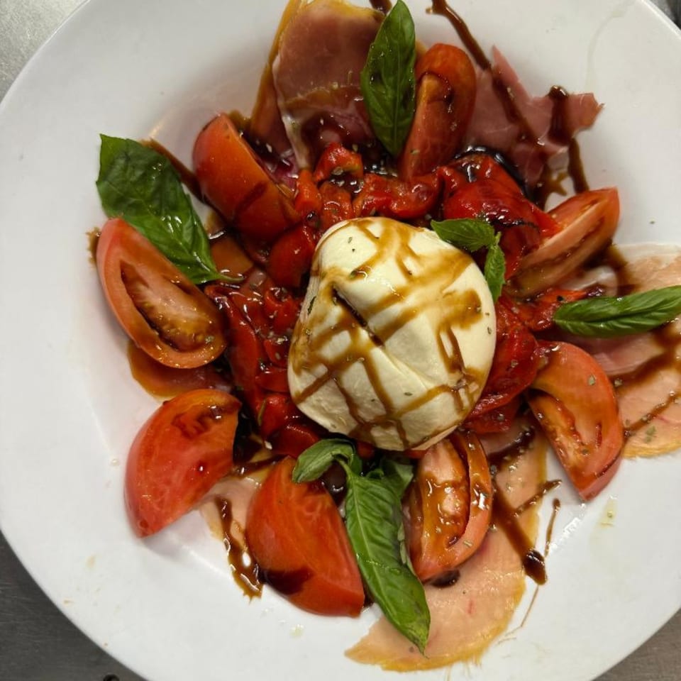 A white plate of Caprese salad with burrata, tomatoes, basil, prosciutto, and balsamic glaze.